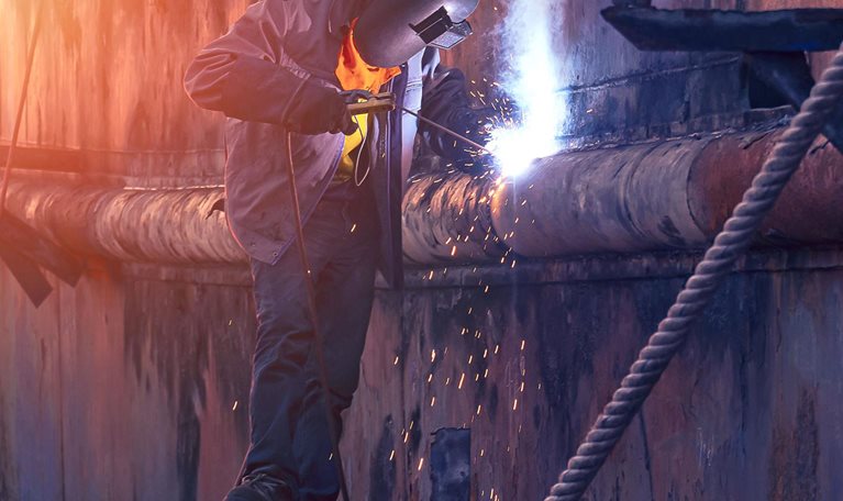 Welder in protective workwear on scaffold is welding the old rusty vessel hull in shipyard area at sunset time