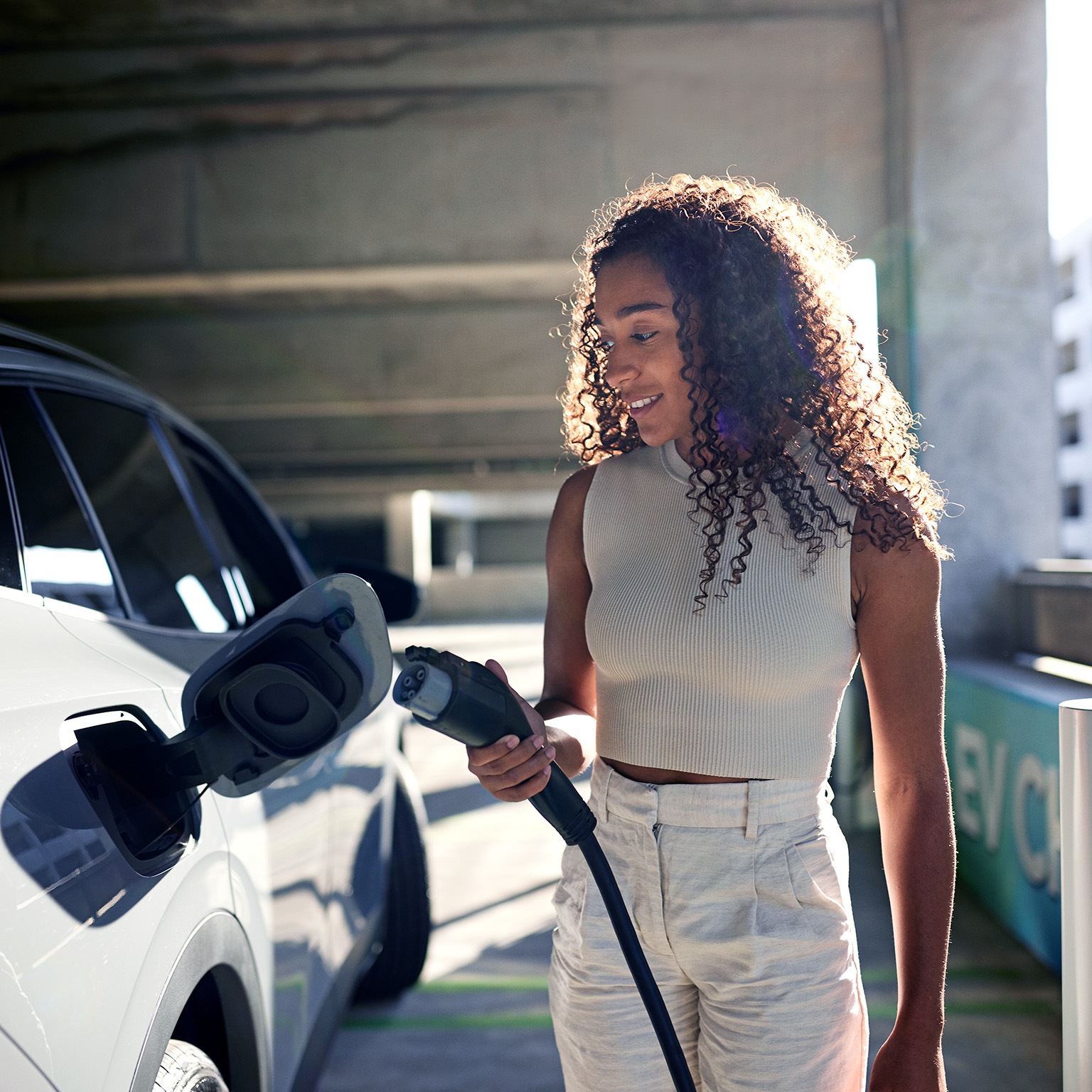 Young woman holding electric plug by car at charging station