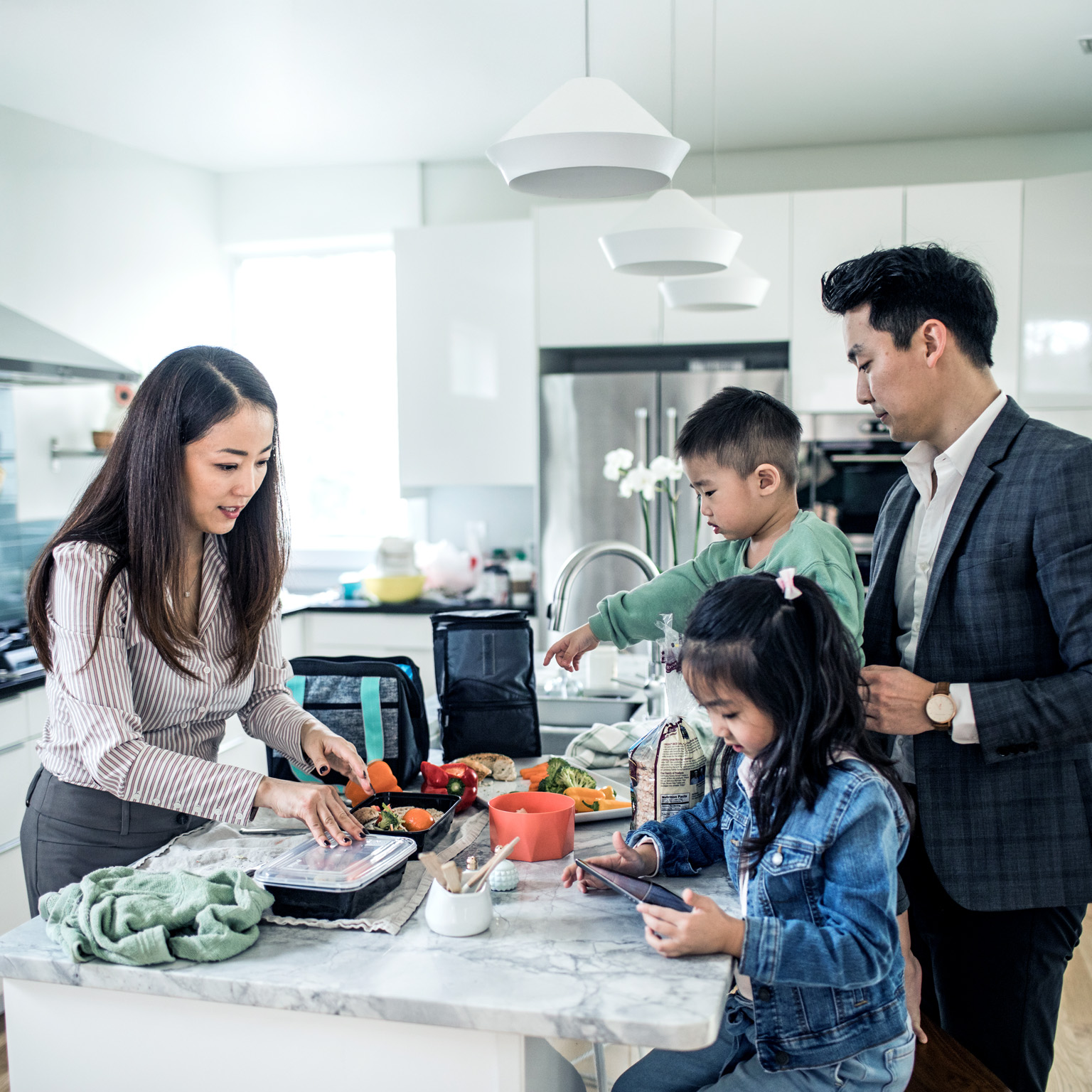 A family gathered in a bright modern kitchen, with parents and two children preparing food and packing containers together. 