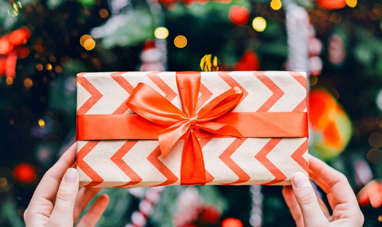 A pair of hands holding a wrapped gift box decorated with a red chevron pattern and tied with a bright red ribbon. In the background, colorful lights and ornaments create a festive, holiday atmosphere.