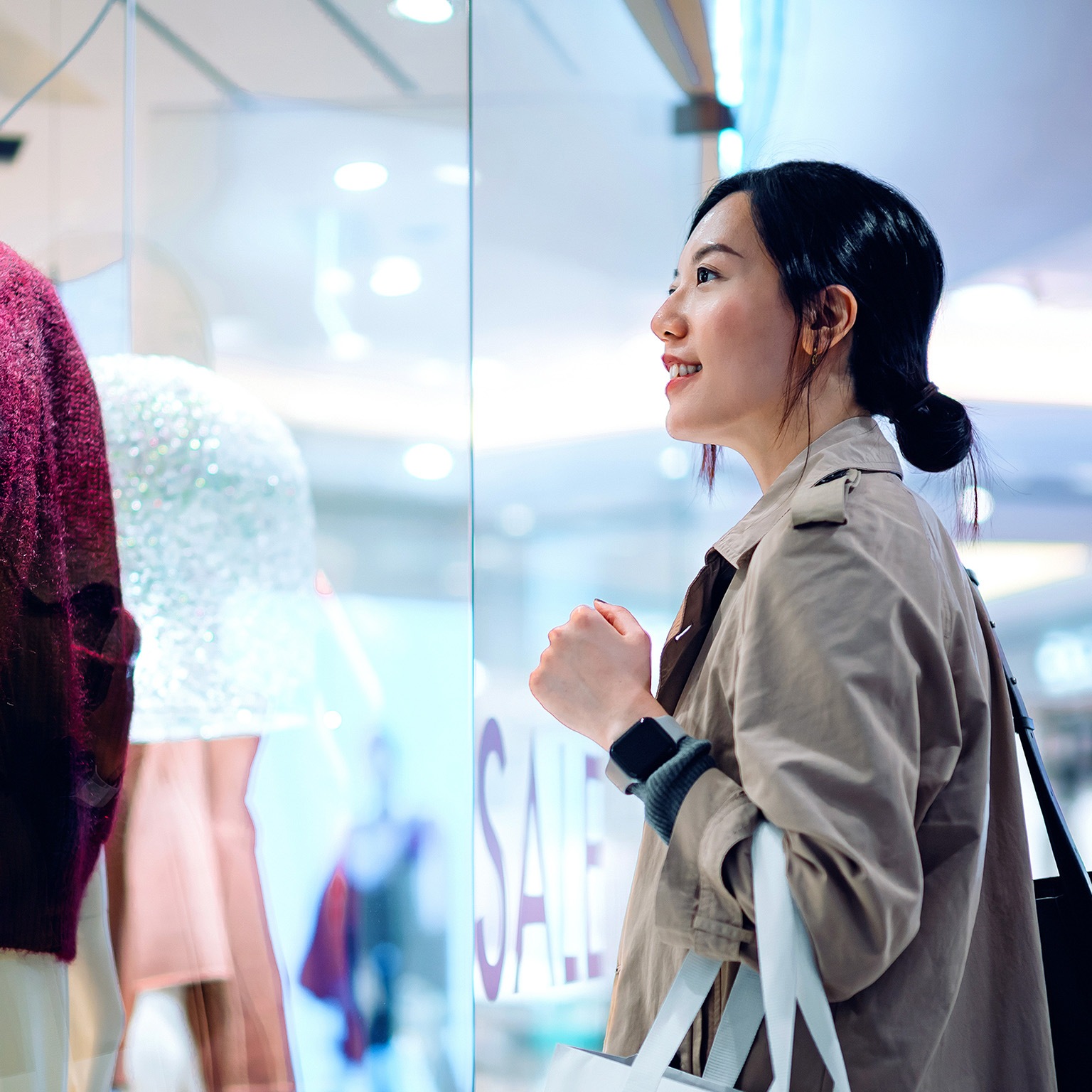 Asian woman carrying a shopping bag, standing outside a boutique looking at window display in a shopping mall