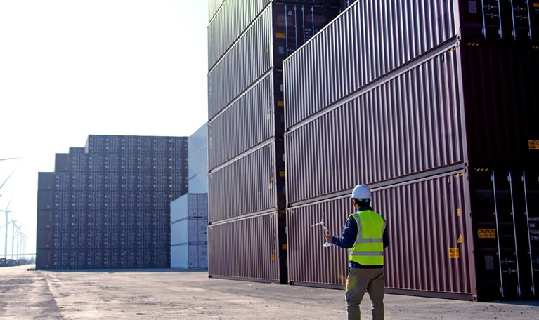 A worker in a dockyard, donning a helmet and reflective vest, gazes at a blueprint of a windmill. In the distance, there are rows of trucks and wind turbines.