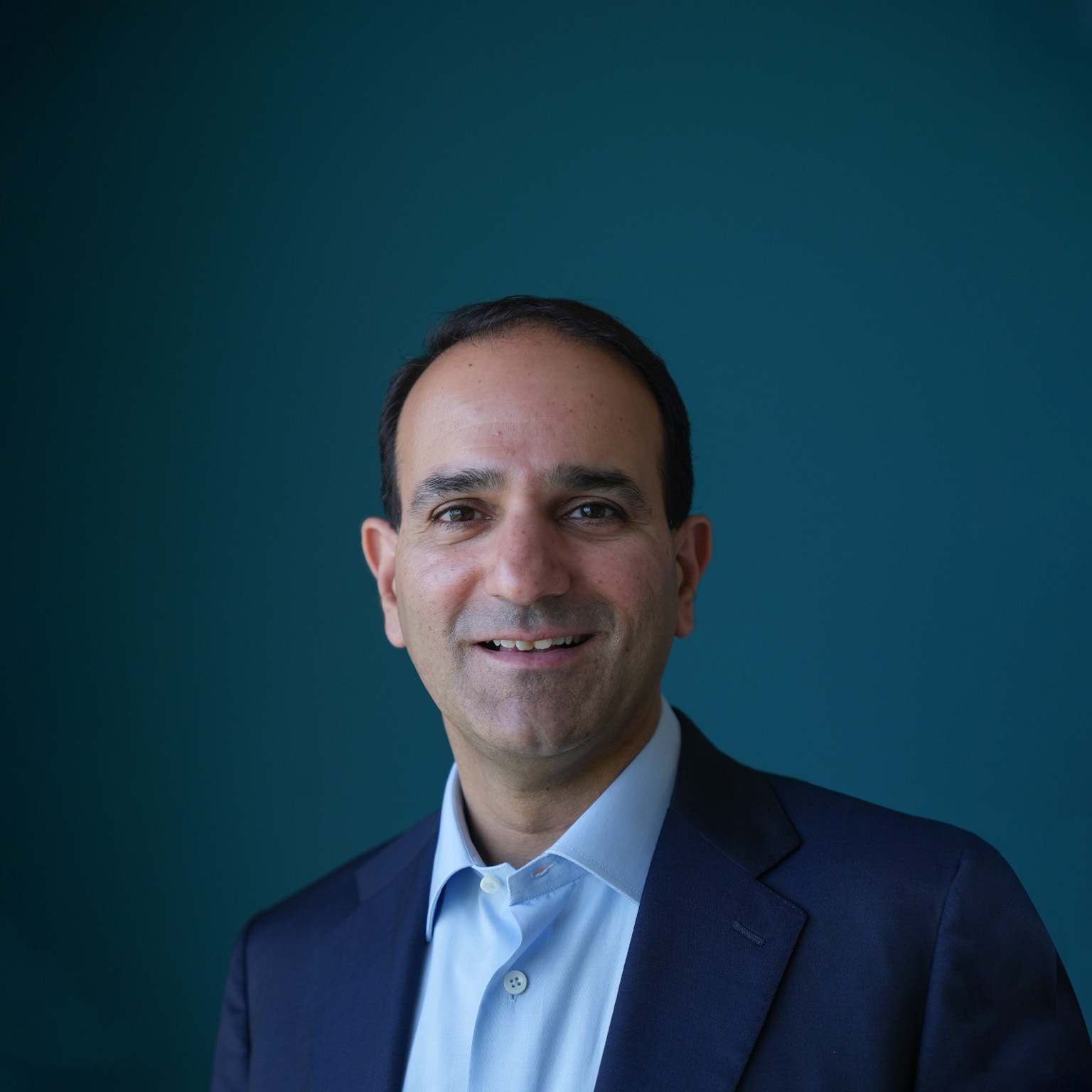Anant Bhalla wearing a dark suit and a blue dress shirt. His face bears a light smile as he poses against a blue backdrop.