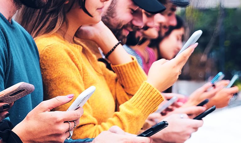 Closeup image of a group of interracial gen-z friends sitting outdoors in a row, looking at and interacting with mobile phones.