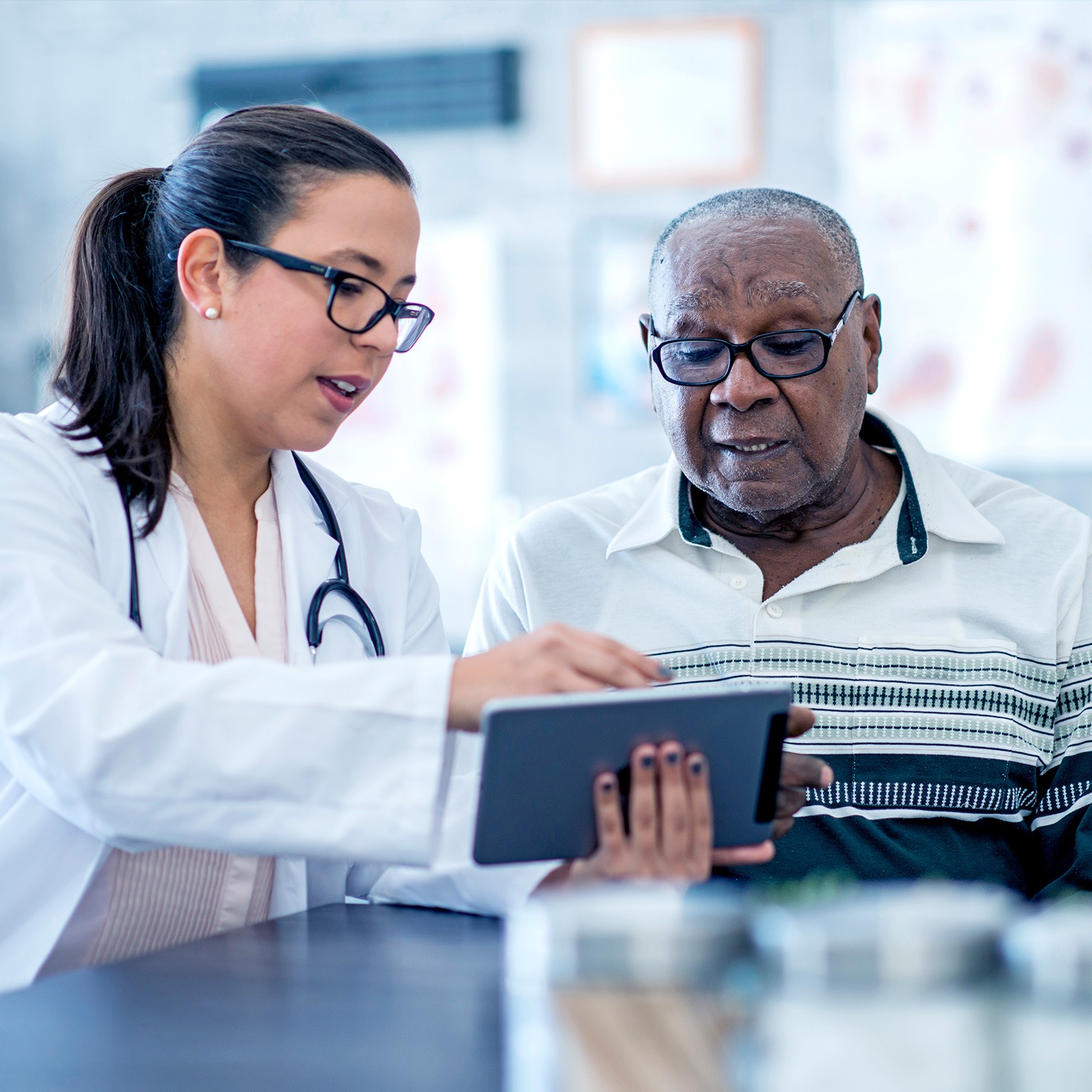 A doctor in white coat wearing a stethoscope, who is a woman, appears to explain something from an electronic tablet to an elderly black man.