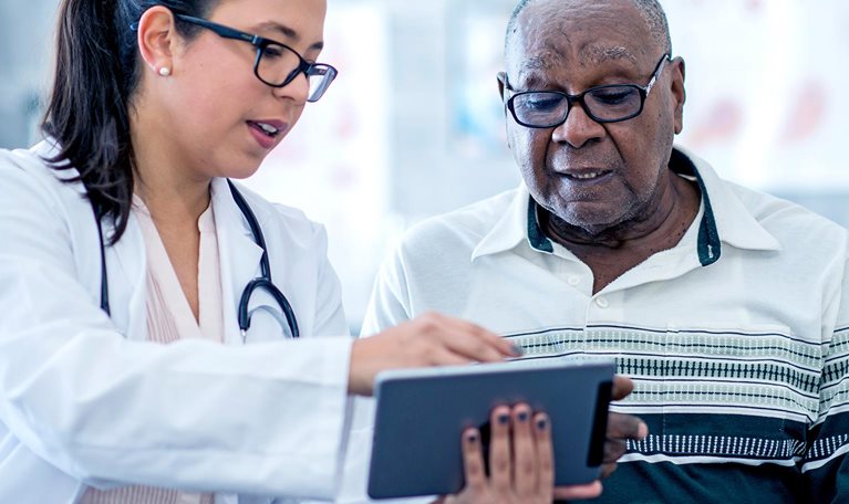 A doctor in white coat wearing a stethoscope, who is a woman, appears to explain something from an electronic tablet to an elderly black man.