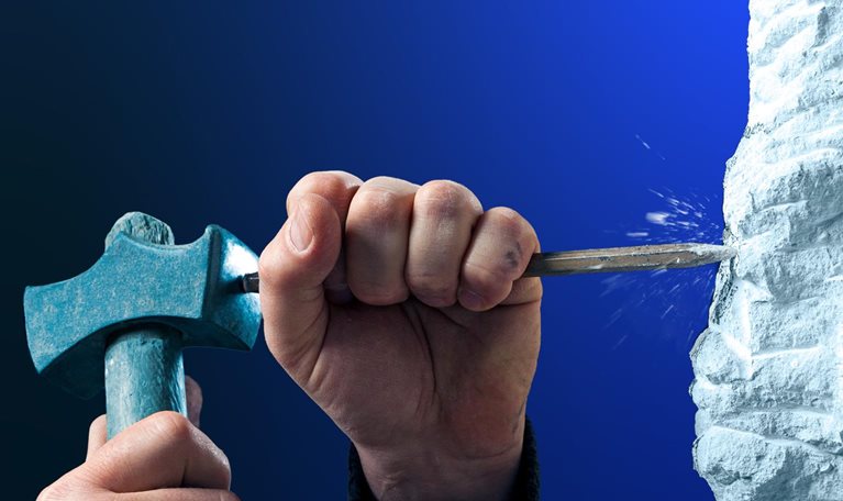 Two hands using a hammer and chisel to carve into a rough white stone surface. The close-up composition is set against a deep blue background.