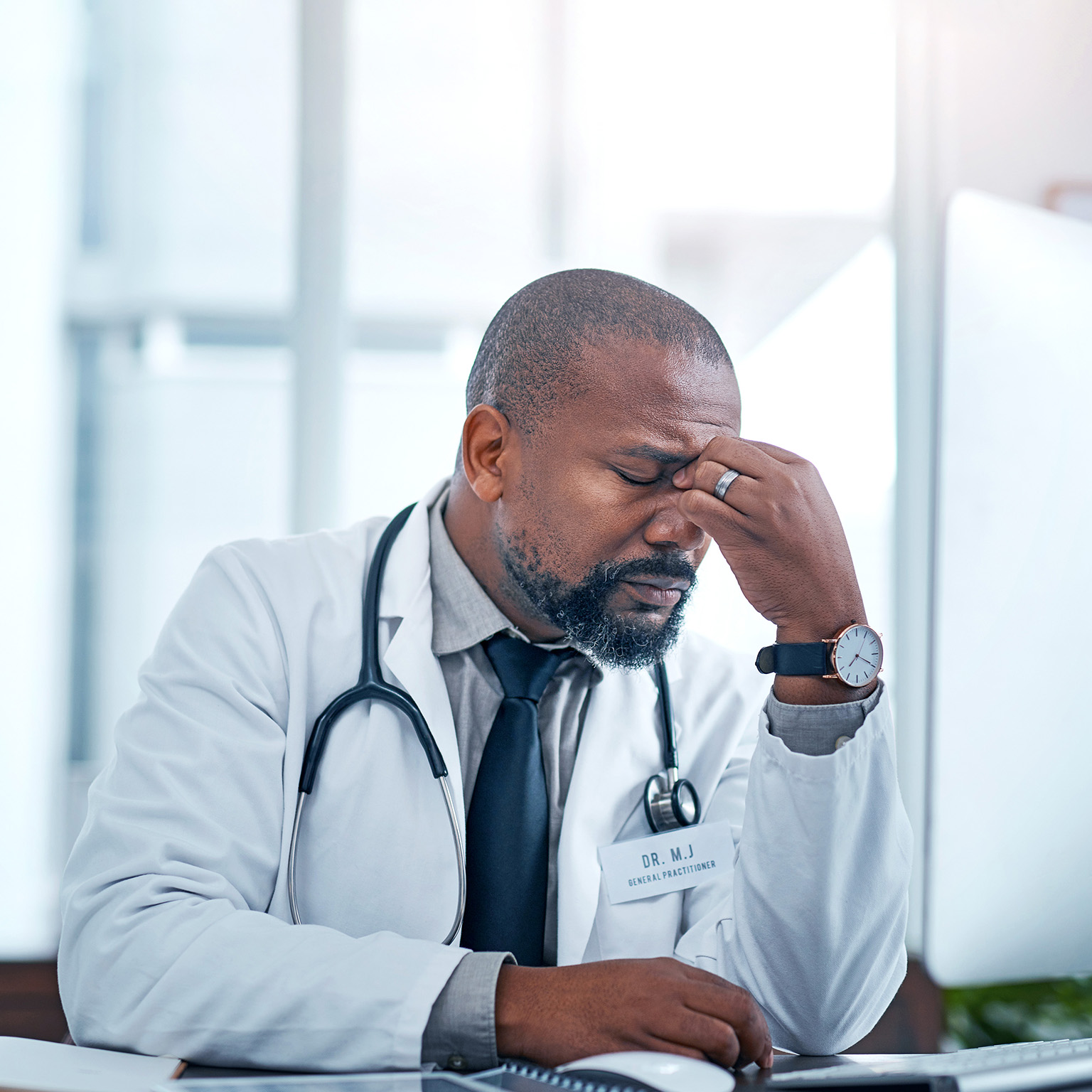 A Black male doctor sits at a computer looking frustrated