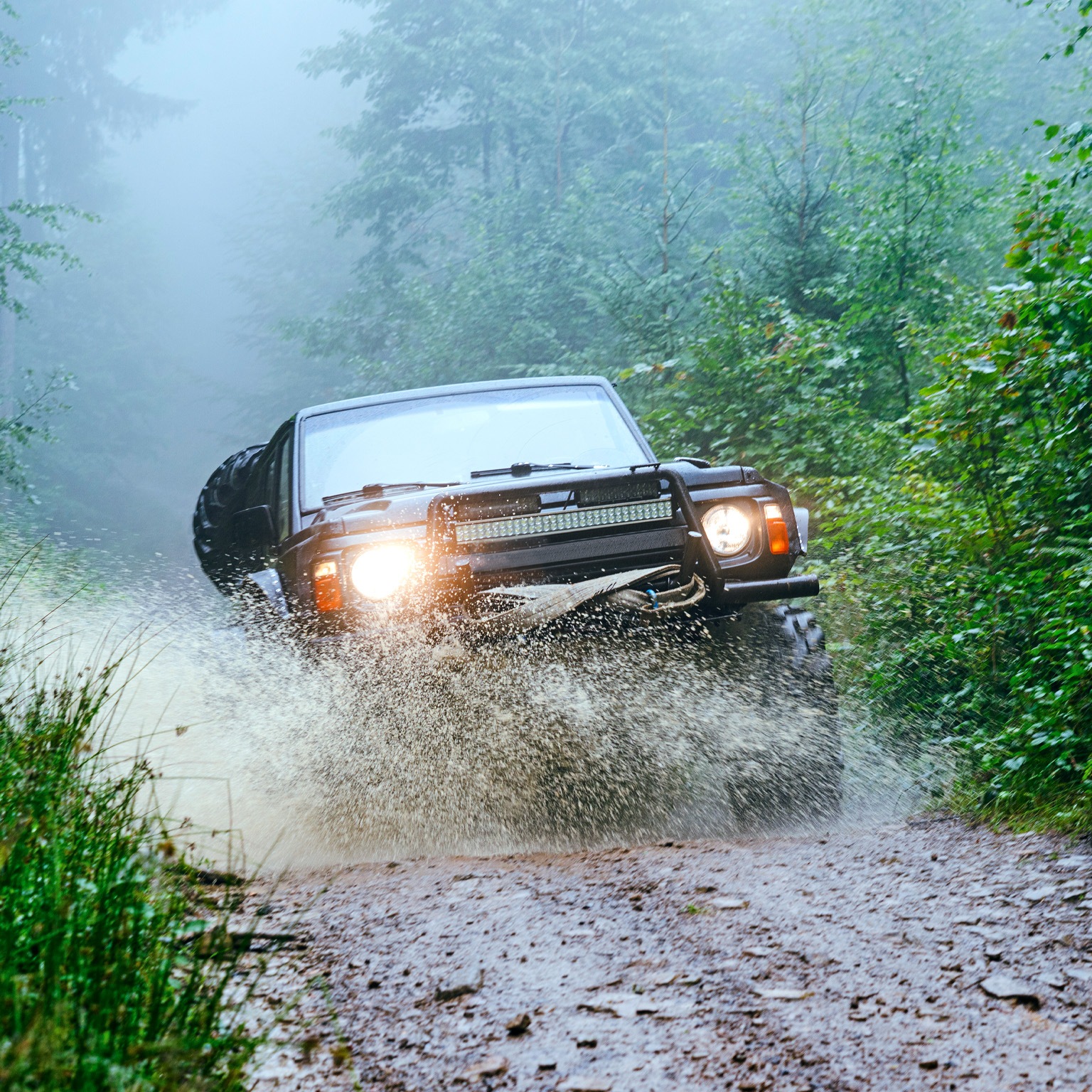 The image shows a rugged off-road vehicle driving through a muddy forest trail, splashing water as it moves forward. Dense green foliage surrounds the path, and mist hangs in the air, creating a dramatic and adventurous atmosphere.