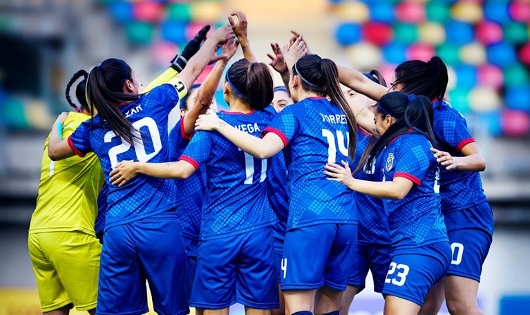 A professional women's soccer team forms a circle with their arms raised and embracing.
