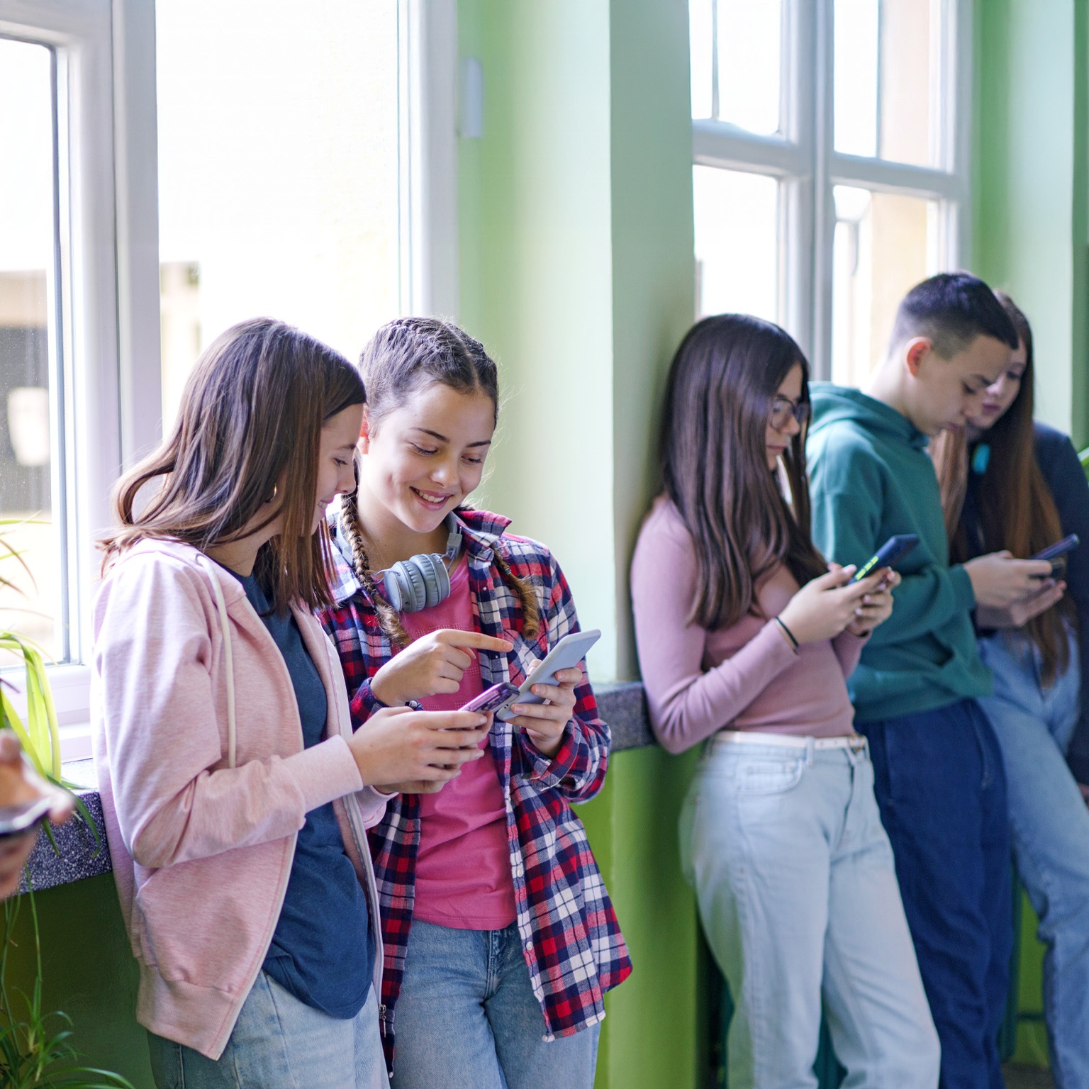 A group of teenagers are standing against a wall, looking at their smartphones. One of the girls in the middle is pointing at her phone, sharing something with another girl beside her.