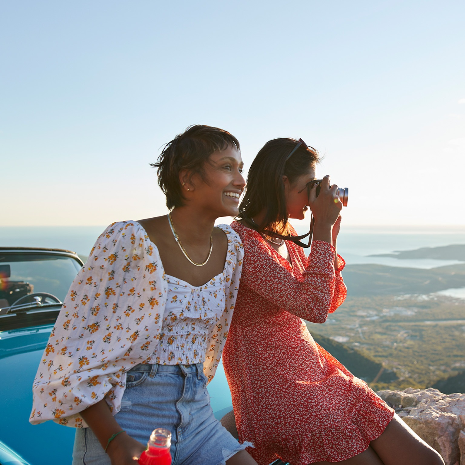 Close up image of friends on vacation sitting on the hood of a car smiling and taking pictures of the view in Montenegro.
