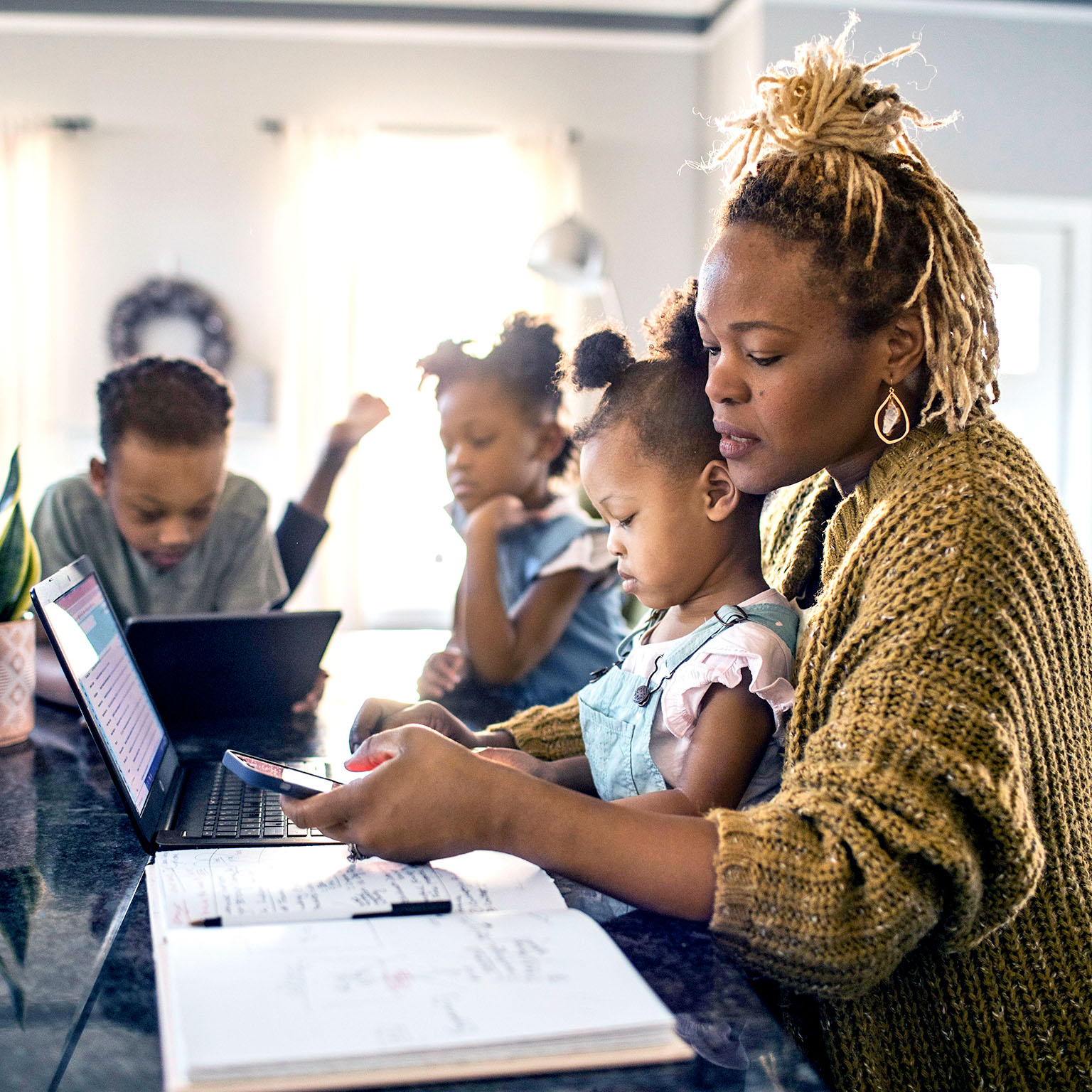 Mother working from home while holding toddler, family in background - stock photo