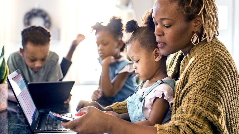 Mother working from home while holding toddler, family in background - stock photo