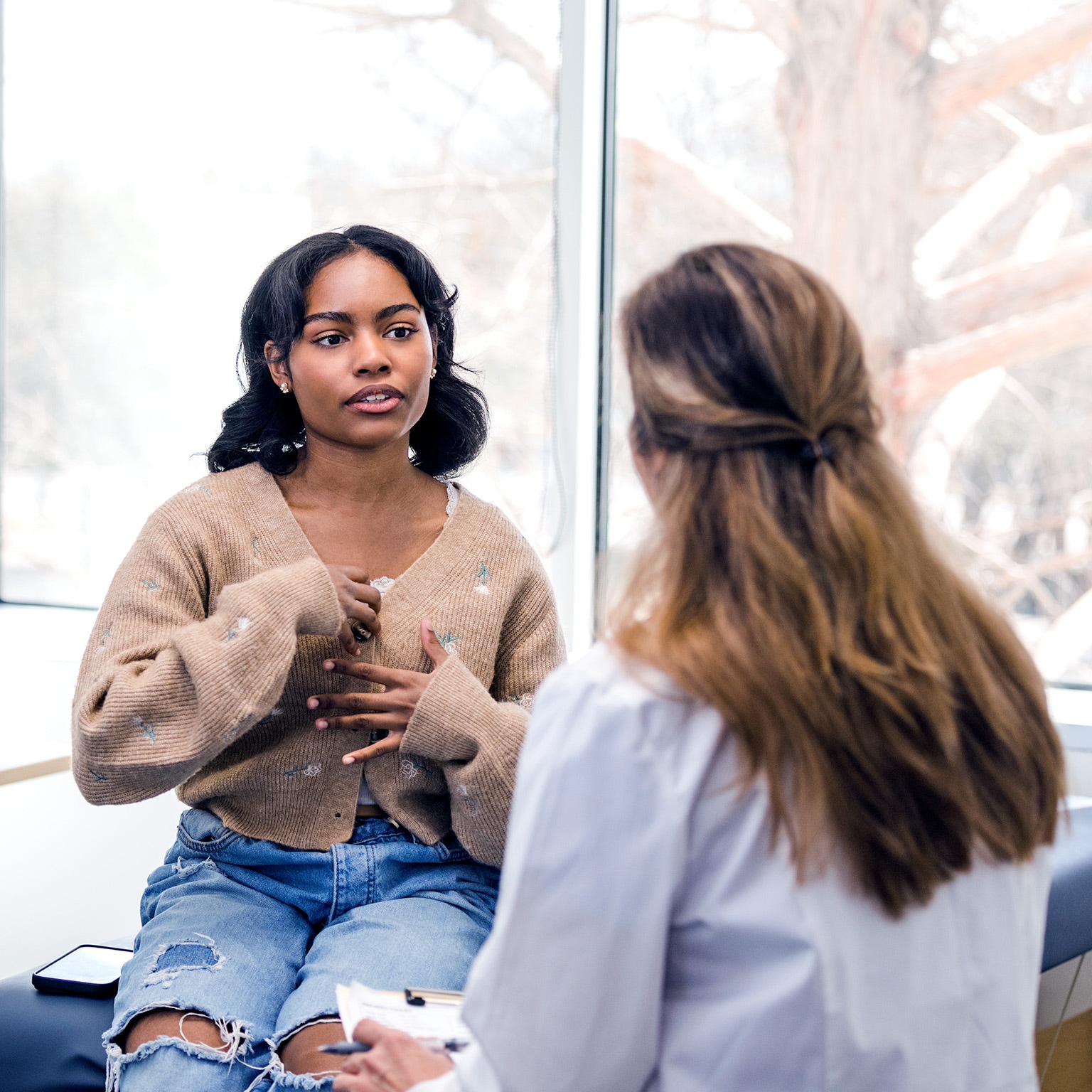 The young adult woman gestures as she explains her symptoms to the unrecognizable female doctor.