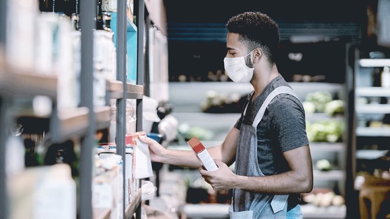 Man working at a supermarket restocking the shelves and wearing a facemask - stock photo