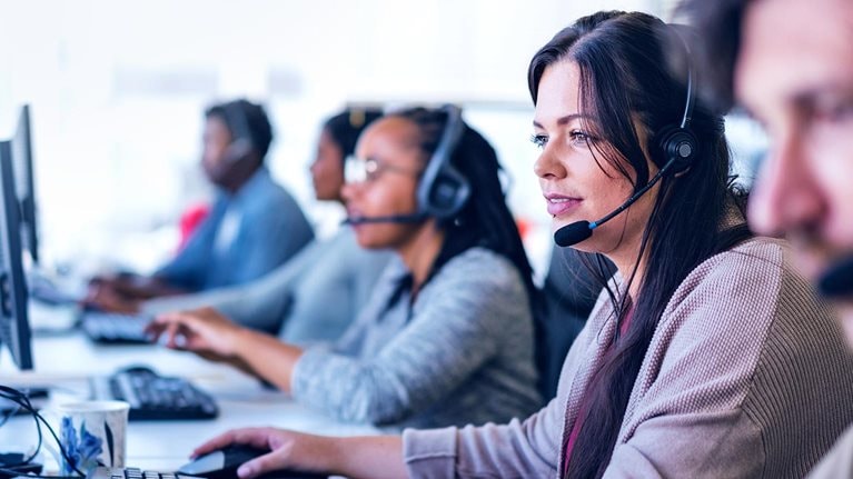 Young businesswoman wearing headset while using computer. Row of operators are sitting at desk. They are working in call center. - stock photo