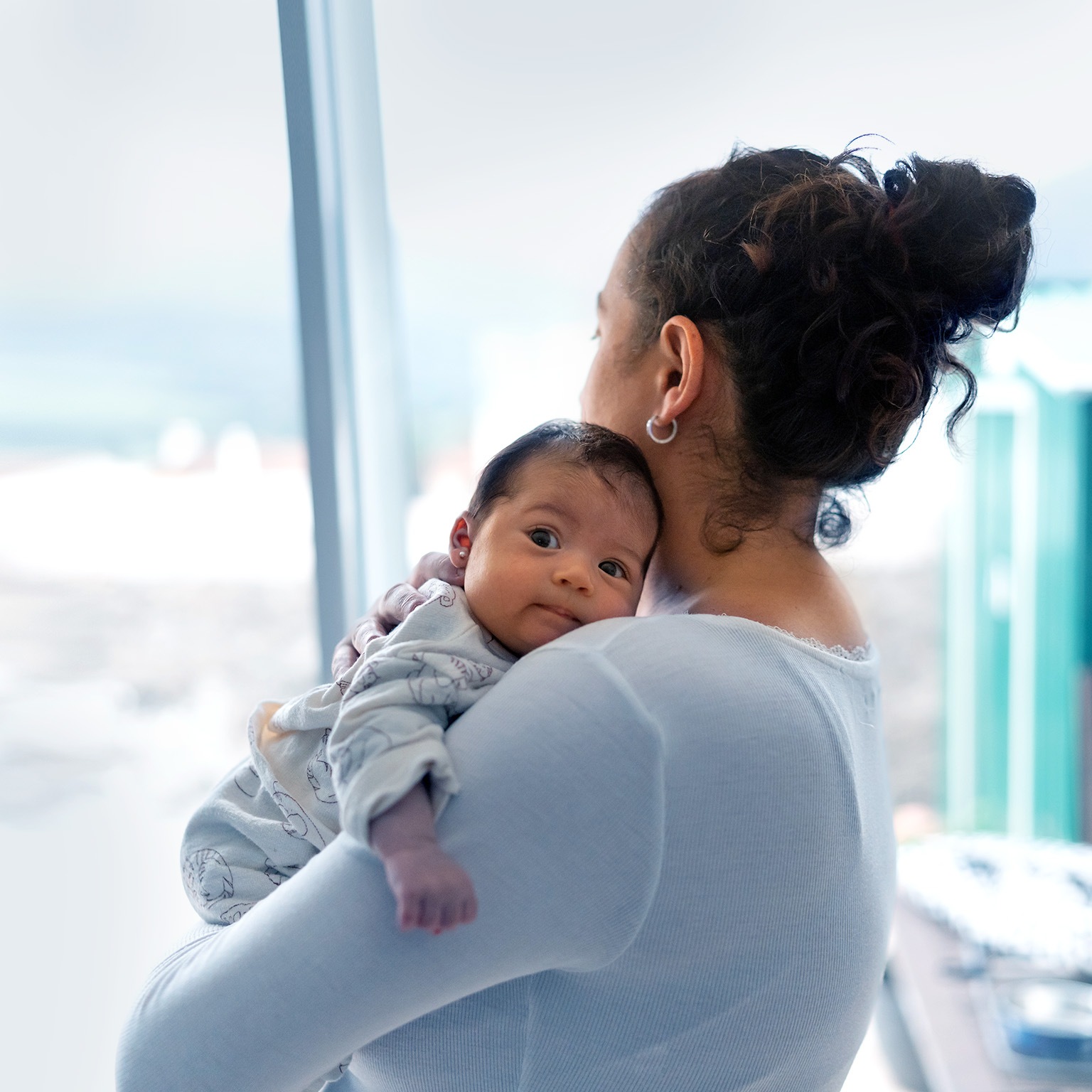 The mother is on her back looking out the window while holding the baby in her arms. The baby is looking at the camera. Concept of protection. - stock photo