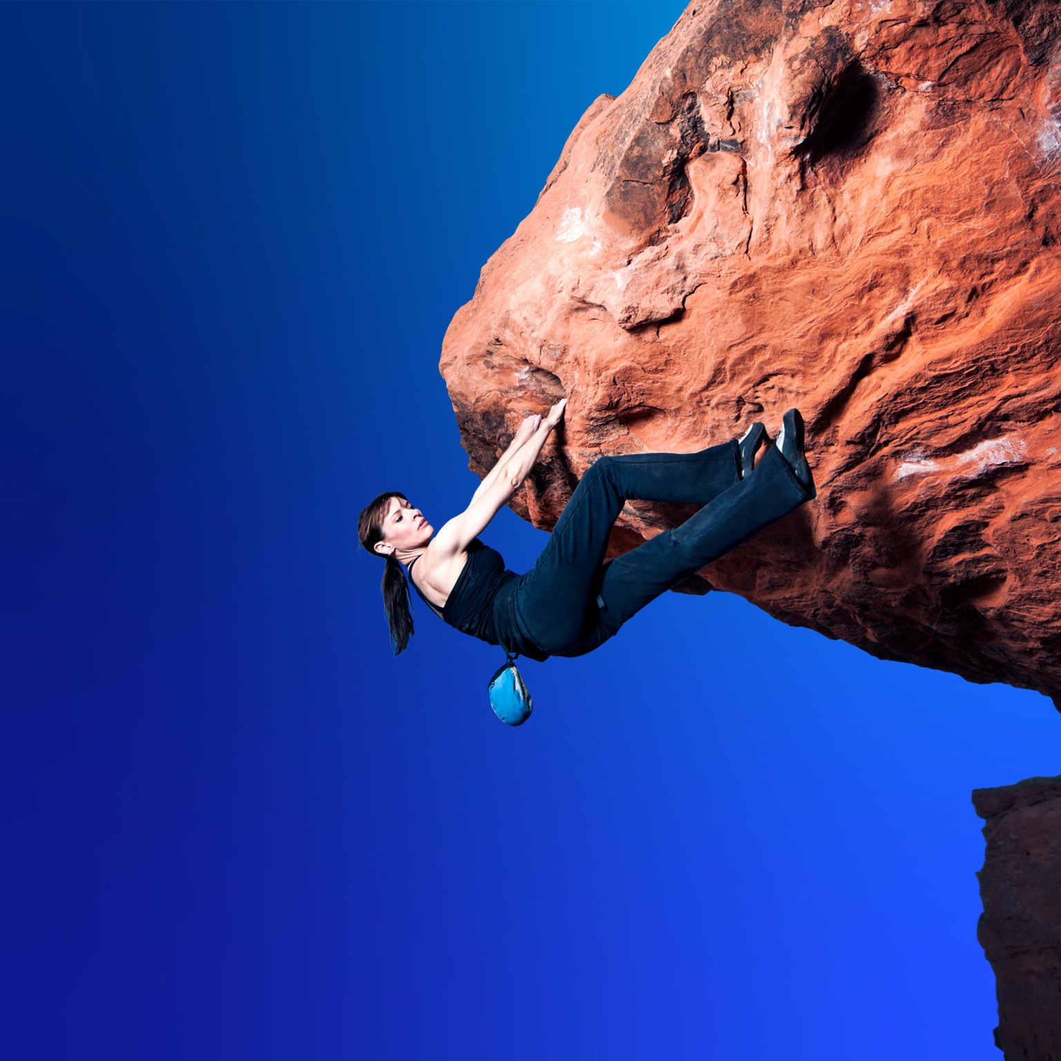 A woman in black rock-climbing clothing navigates a large overhanging bolder without a rope. She has a firm hold with both hands but seeks a foot hold as she makes her way over the protruding rock.
