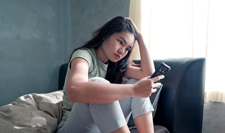 A teenager girl sitting on a day bed and looking at a phone with a concerned expression.