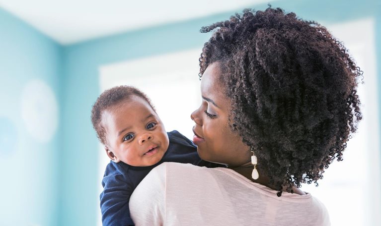 A Black mother with tightly curled ringlets holds her baby on her shoulder. The baby gazes at the viewer inquisitively as the mother looks at him thoughtfully.