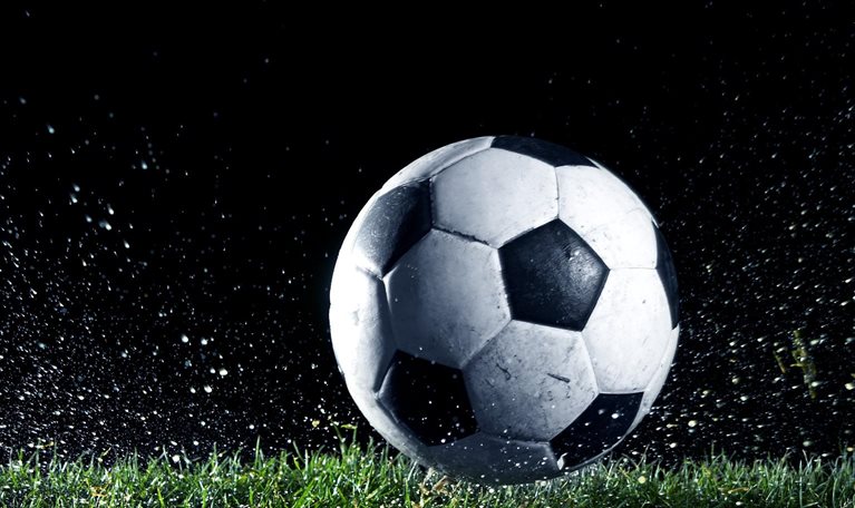 A black-and-white soccer ball sits on wet grass as water and turf spray outward, suggesting recent impact or motion.
