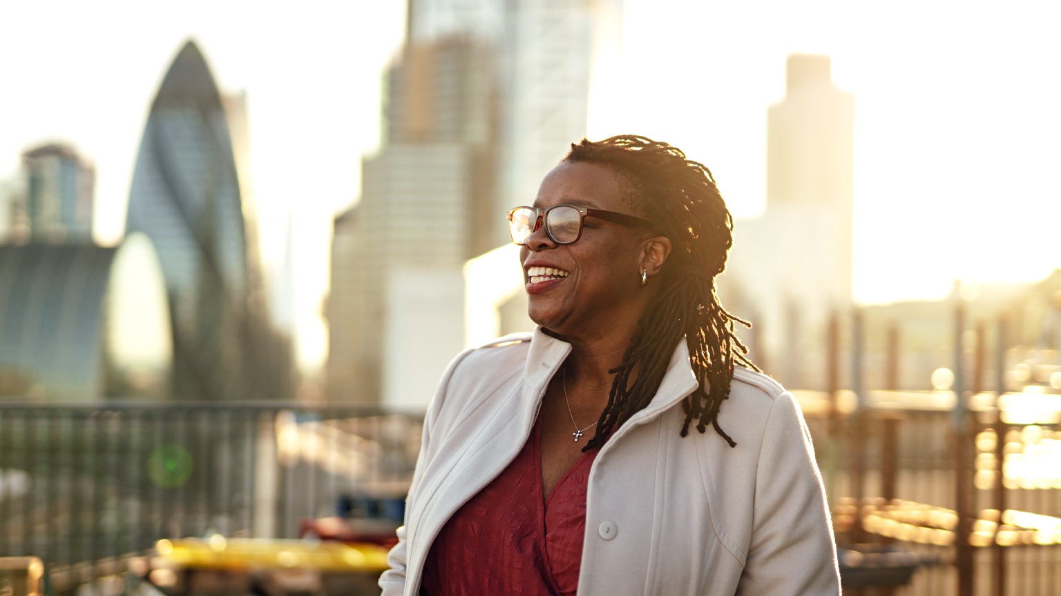 A mature businesswoman standing with her hands in pockets on office building rooftop with the London cityscape in the background. 