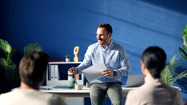A cheerful CEO stands beside his desk, attentively hearing out a diverse group of employees seated in chairs.