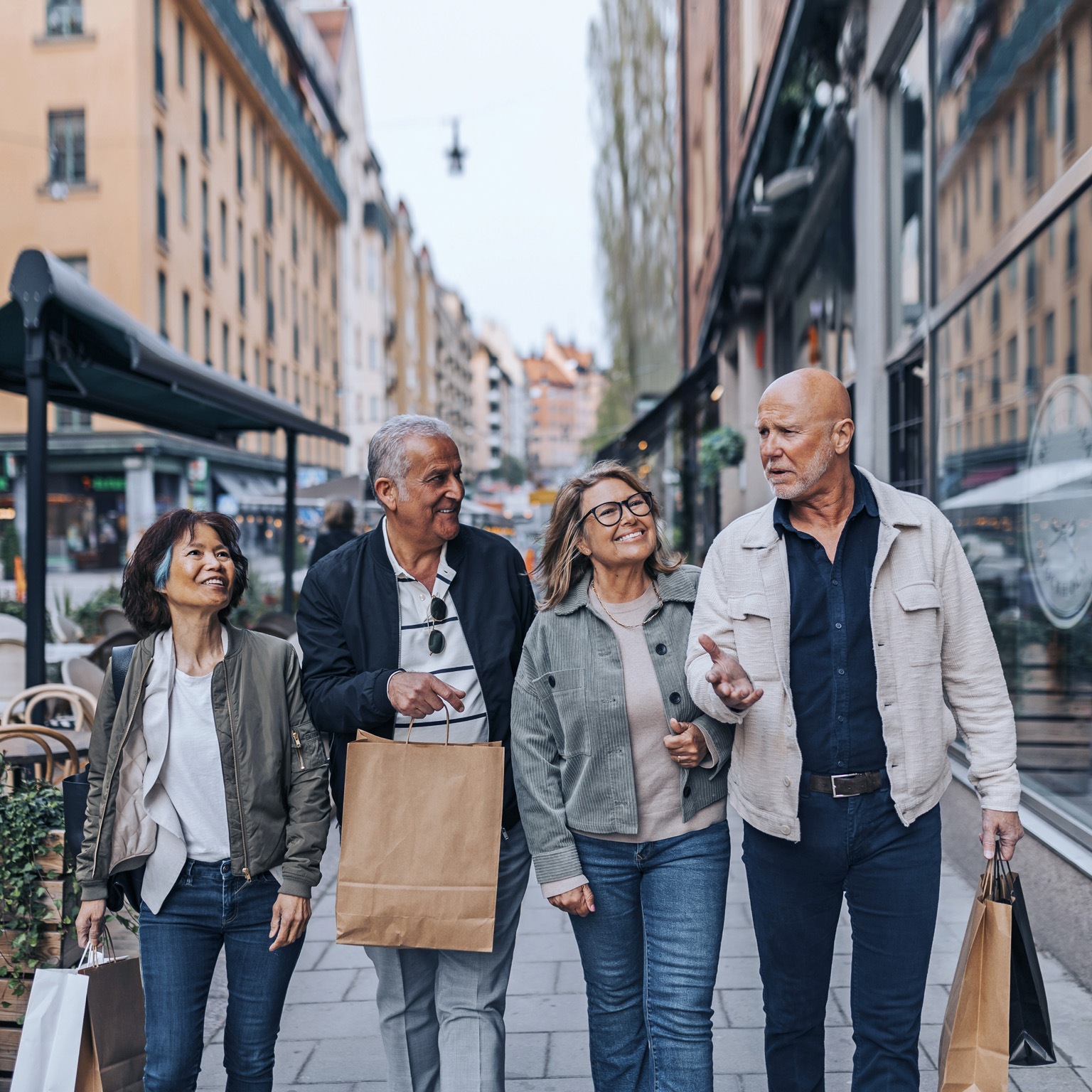 A cheerful group of senior friends strolling down a European sidewalk, engaged in conversation and carrying shopping bags.