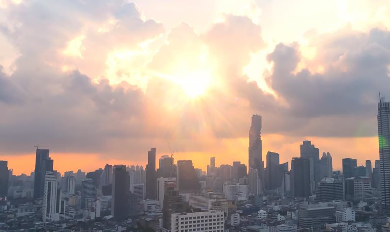 The image is an aerial view of the Bangkok skyline, likely near the Sathorn district, featuring several modern skyscrapers at sunset or sunrise. The sun is low on the horizon, casting a warm orange glow and dramatic light rays through the clouds.