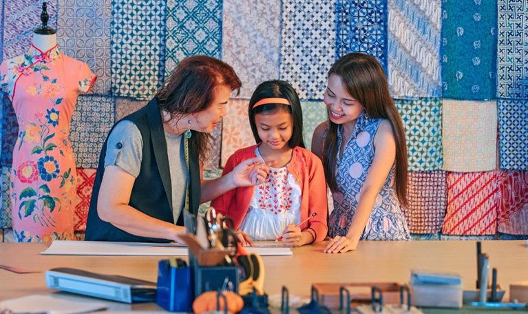 A Chinese grandmother teaching her granddaughter how to design a garment while her mother looks on. Fabric patterns hang on the wall behind them and traditional Chinese dresses hang on mannequins.