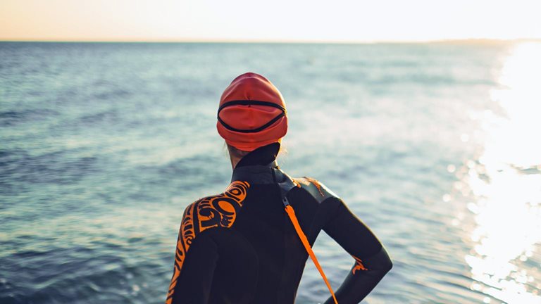 A triathlete wearing a wetsuit and swim cap stands facing a calm ocean. Her back is to the viewer as she takes in the setting sun. 