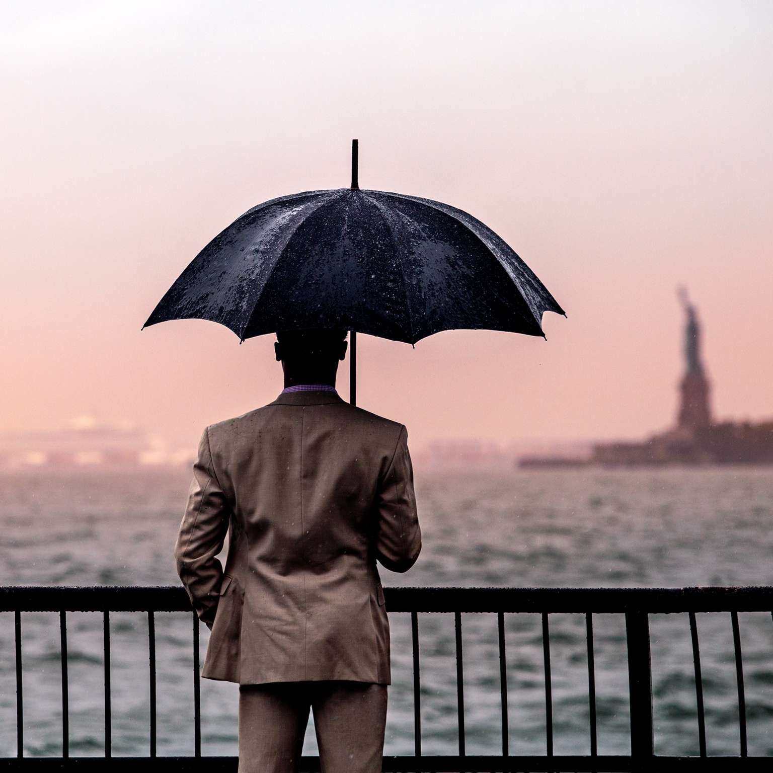 Image of a man looking at the Statue of Liberty in the distance while holding an umbrella