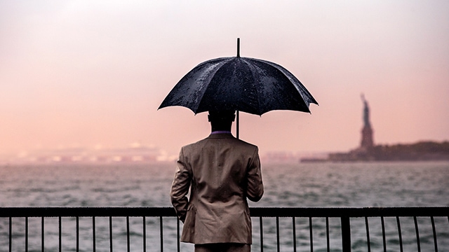 Image of a man holding an umbrella and looking out across New York Harbor to the Statue of Liberty