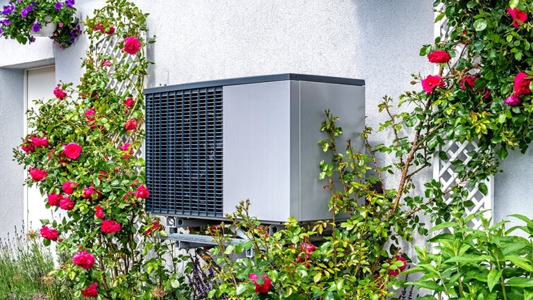 Image of a heat pump attached to a house with flowers growing around it