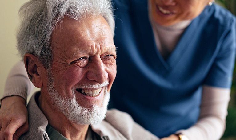 Portrait of a Smiling Elderly Man Taking a Jacket off With a Help of a Woman in a Care Home
