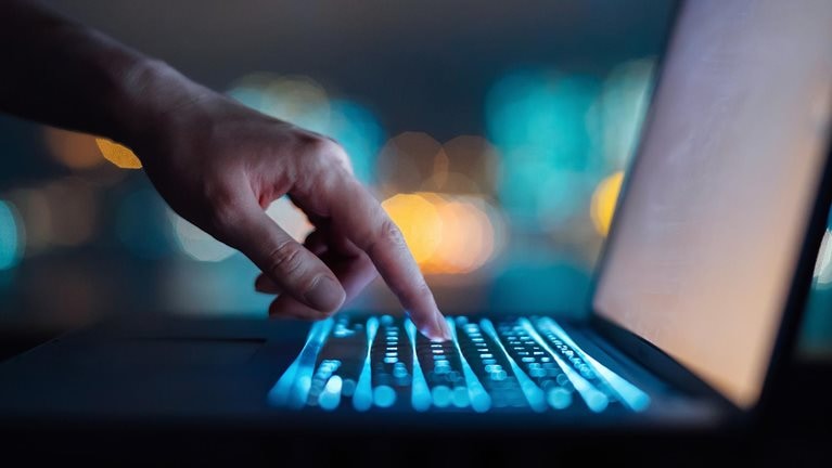 Close up of woman's hand typing on computer keyboard in the dark against colourful bokeh in background, working late on laptop at home
