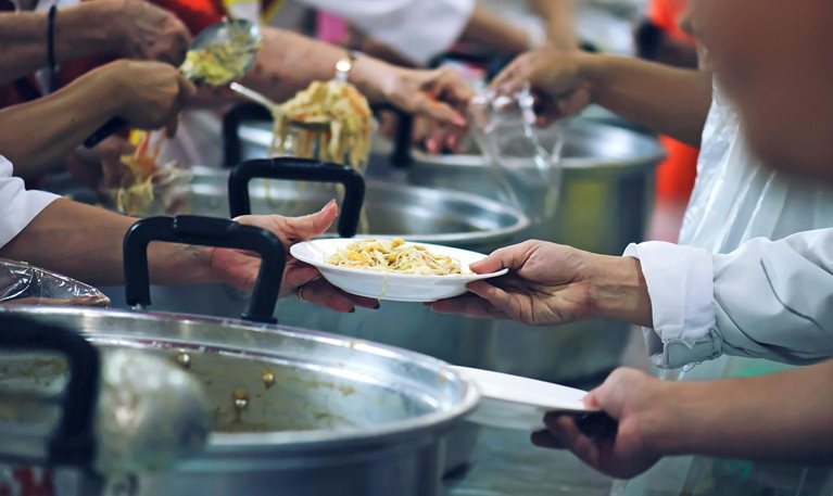 A close-up of several people serving food in a communal setting, with hands passing a white plate of noodles over large metal pots while others scoop servings using ladles.