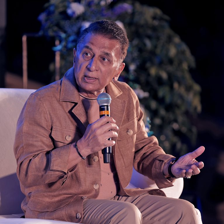 A panel discussion on a stage featuring Sunil Gavaskar seated in a white armchair, holding a microphone and speaking while gesturing with one hand. A leafy plant arrangement is visible in the background against a softly lit, dark backdrop.