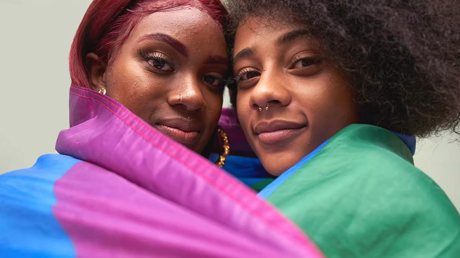 Two women hugging while wrapped in an LGBTQ+ pride flag.