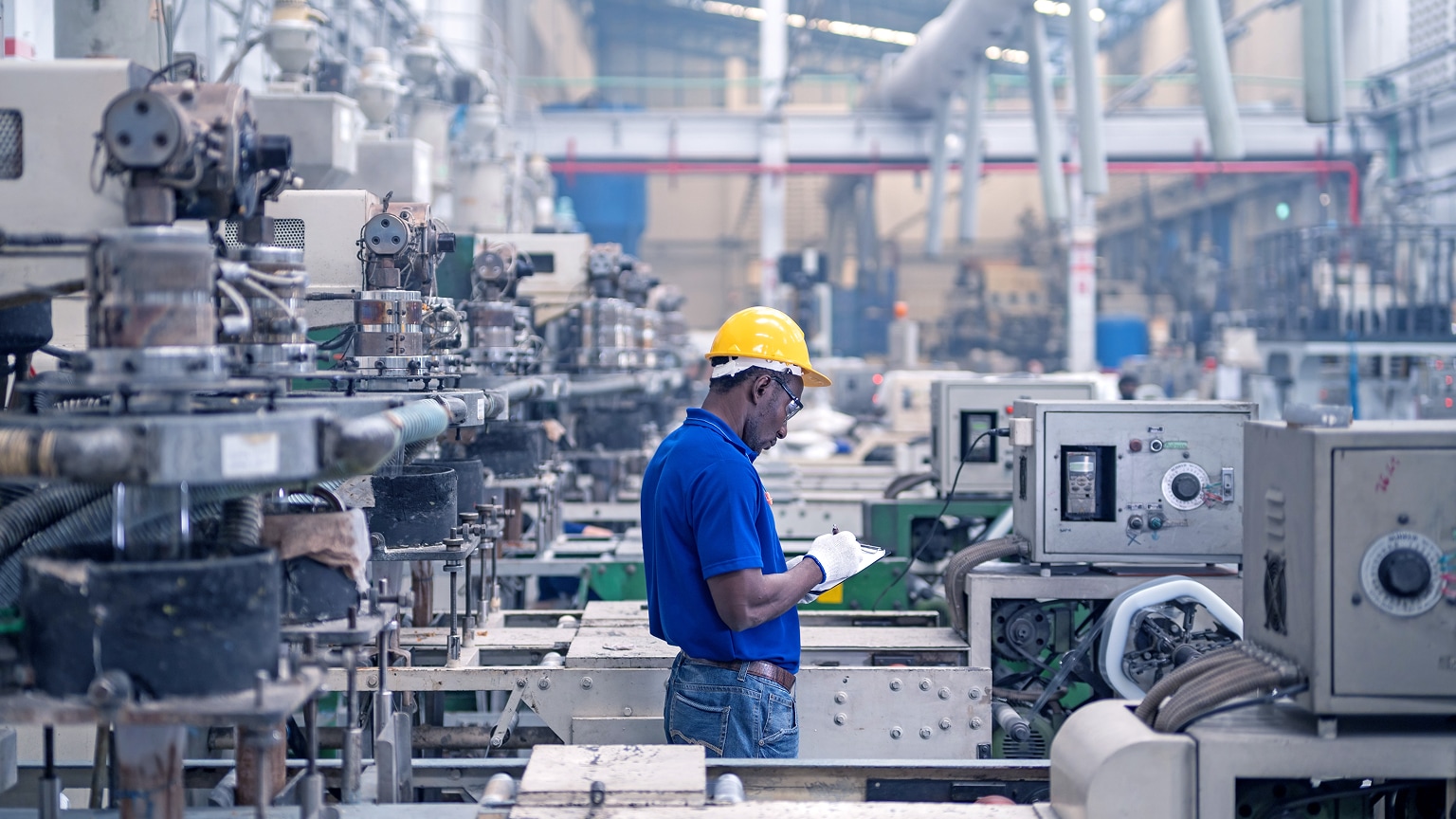 Worker wearing a yellow hard hat records information on a clipboard among industrial machinery in a factory.