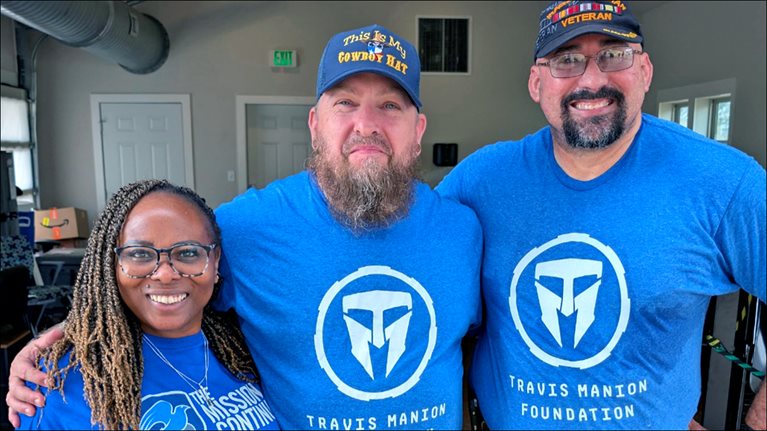 Three people wearing blue Travis Manion Foundation shirts smiling with arms around each other indoors