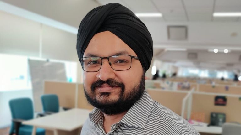 An Asian man wearing a turban and smiling stands in front of an office full of cubicles