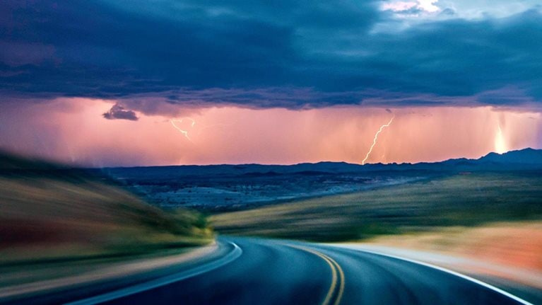 A dark asphalt road curves through a motion blurred landscape beneath a dramatic, stormy sky. In the distance, lightning flashes behind rain clouds, illuminating the horizon with an eerie light.