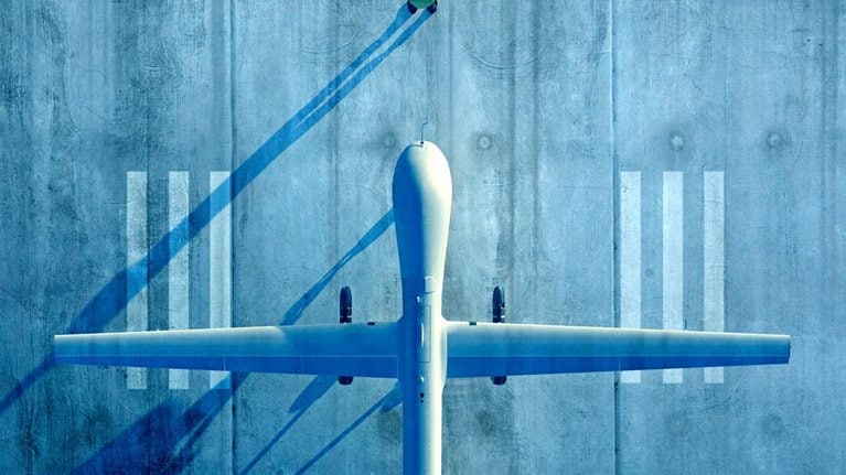 An overhead view shows a large unmanned aircraft parked on a concrete runway, its long wings stretching symmetrically across the frame. A small person nearby casts a long shadow, emphasizing the scale of the scene.