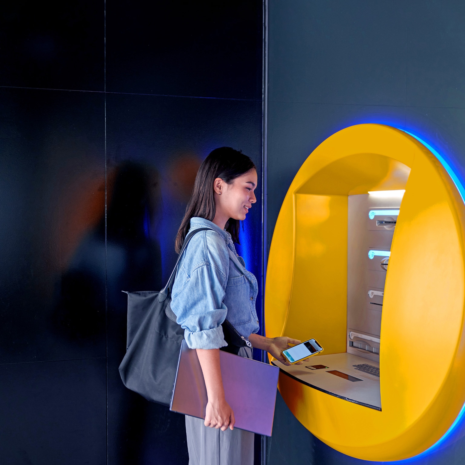 A woman with a smile on her face is standing at an ATM, holding a cell phone and laptop, as she prepares to withdraw money.