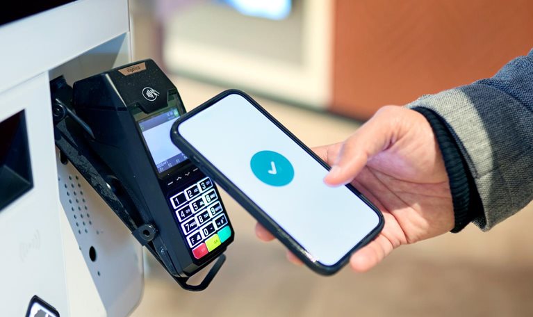 A person holds a smartphone near a payment terminal, with a checkmark displayed on the screen indicating a successful transaction.