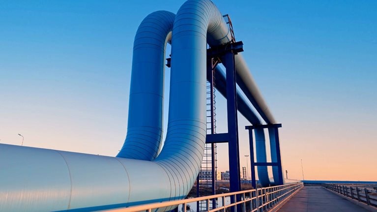 A large industrial pipeline rises and bends over a bridge-like structure, set against a clear blue sky at dusk. A paved walkway with railings runs alongside the pipes, leading the eye toward the horizon and giving the scene a clean, modern, and slightly futuristic feel.