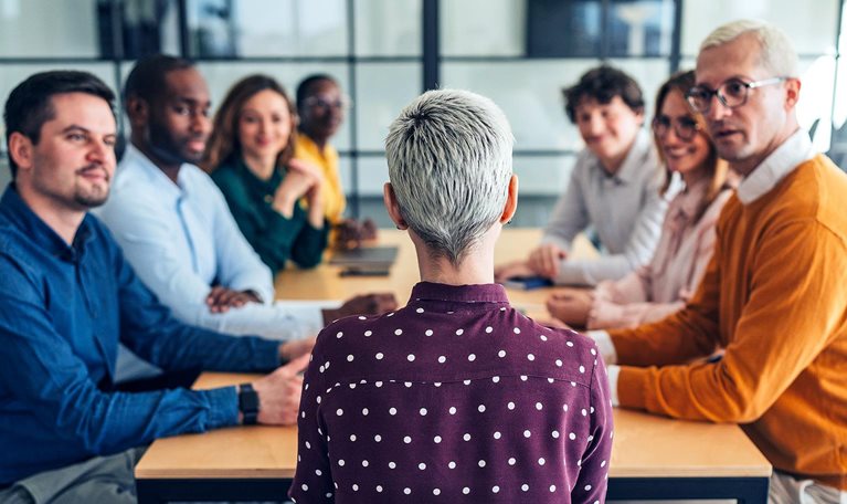 Image from behind of a woman with short gray hair, seated at a conference table, with a diverse group of professionals gathered around, looking at and intently listening to her.