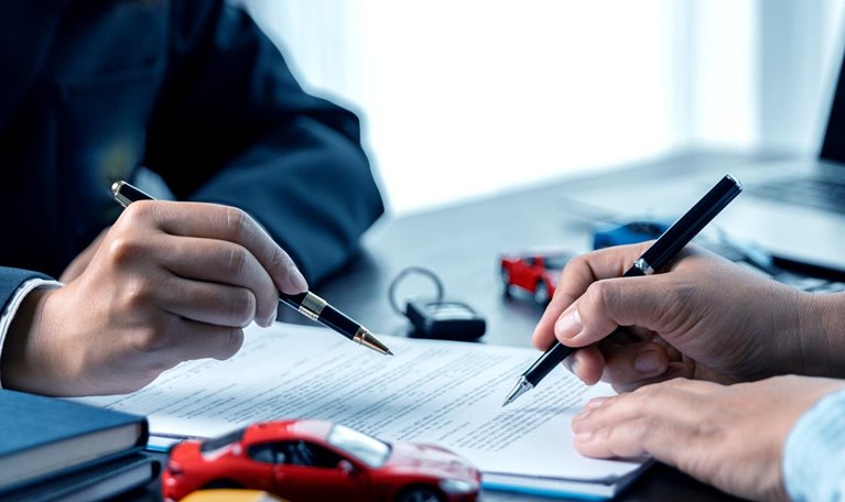Two people signing documents at a desk, with their hands holding pens over a contract. Toy cars and a set of keys on the table suggest the paperwork is related to an auto insurance agreement.